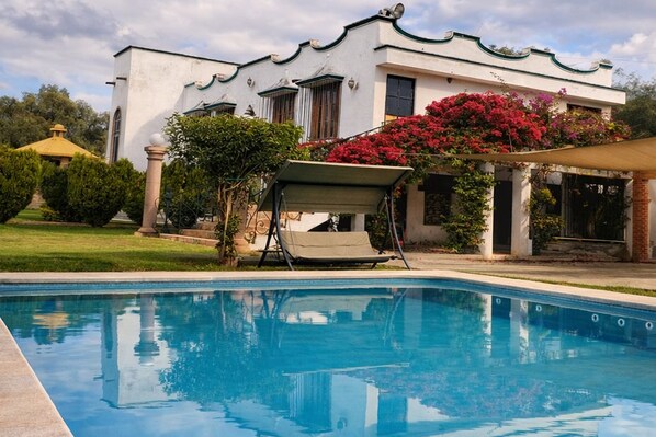 A swimming pool with a white building in the background, a covered seating area, and lush greenery.