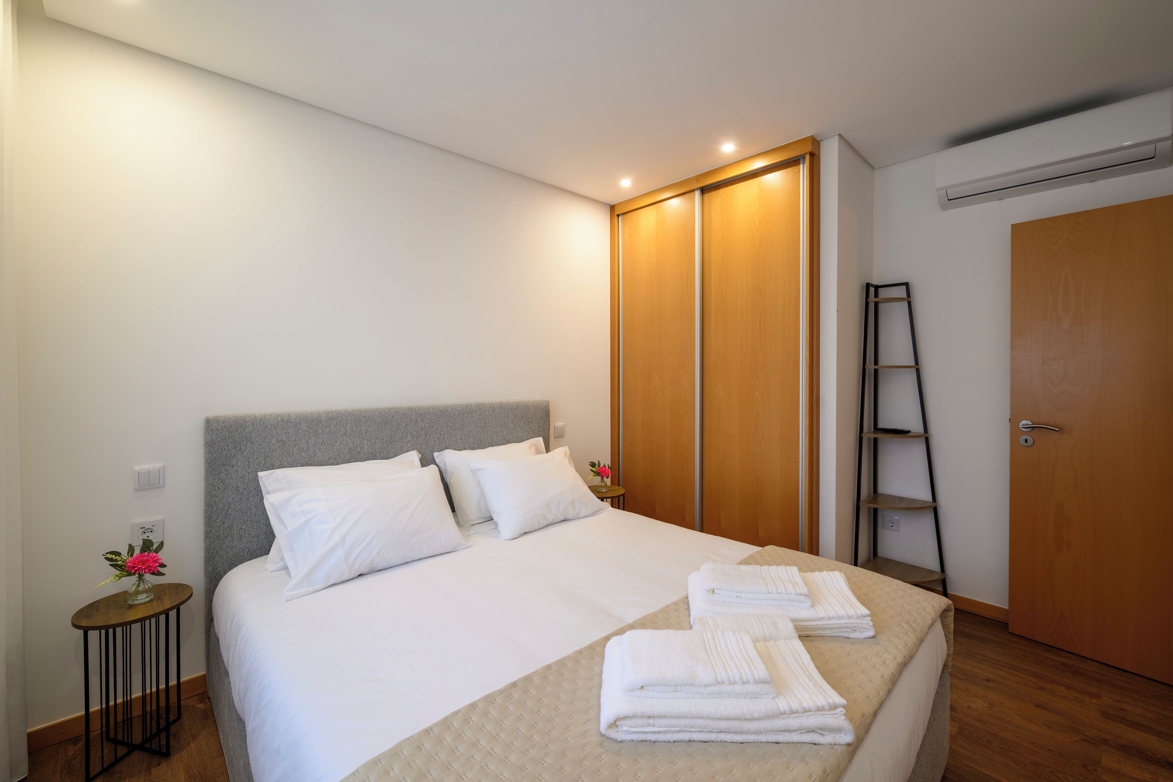 A neatly made bed with white linens and towels, a grey headboard, and a wooden wardrobe with a sliding door.