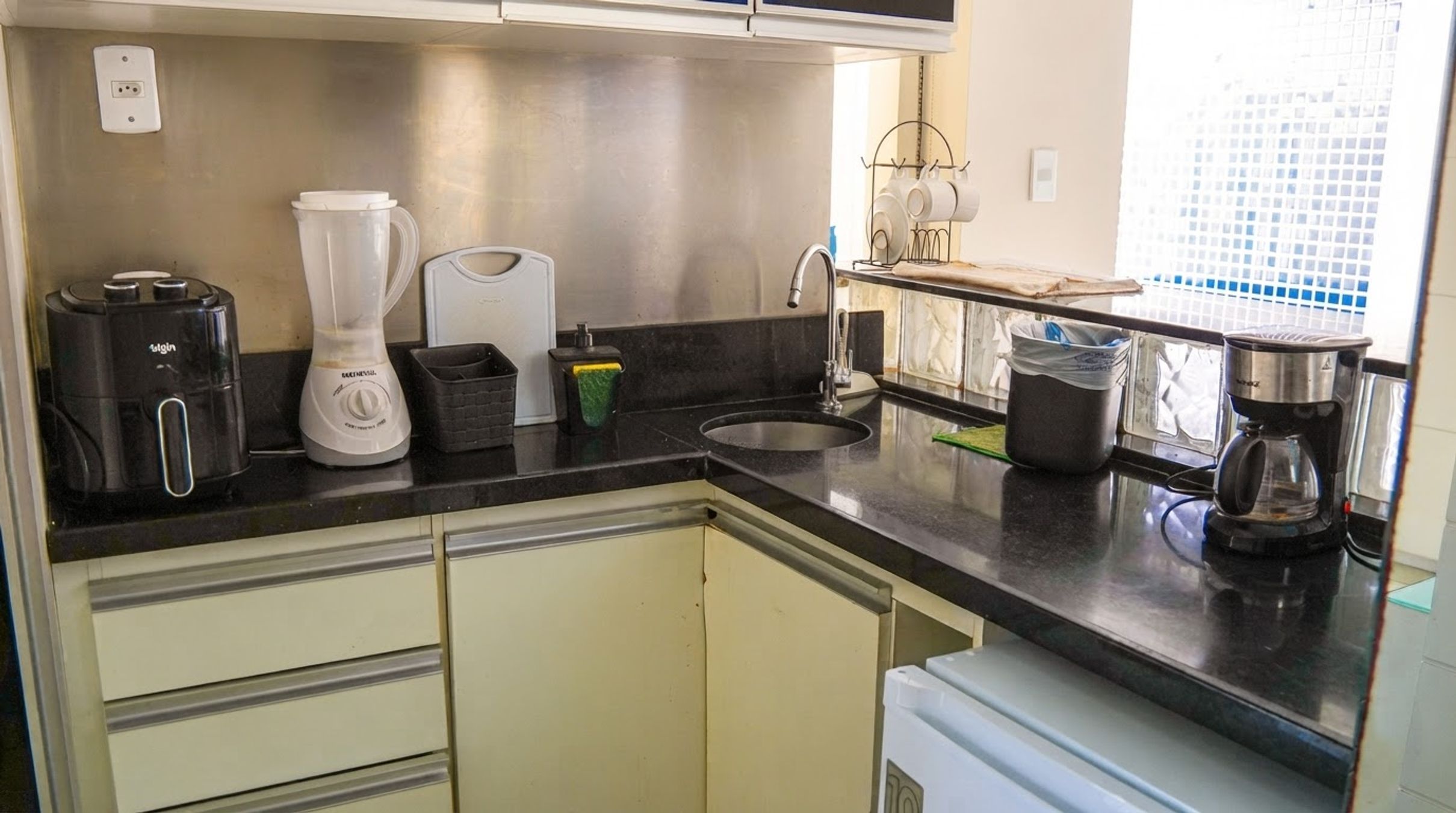 Una cocina compacta con encimera negra, cafetera, licuadora y pileta.