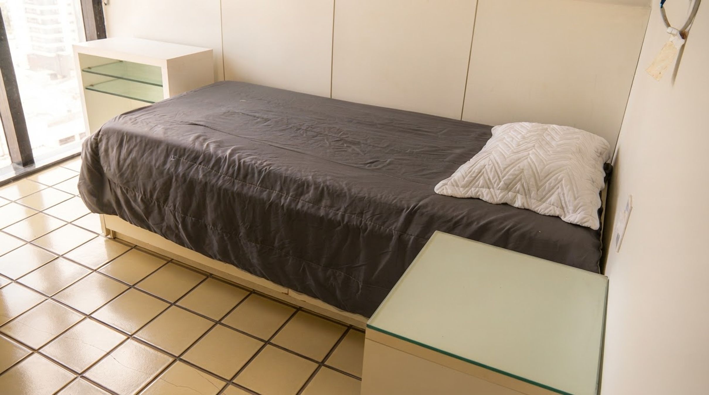Un letto singolo con copriletto scuro e cuscino bianco, un piccolo comodino e un tavolino con piano in vetro.