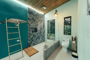 Une salle de bain avec un mur orné d’un motif en chevrons, une échelle en bois, une cabine de douche en verre et des toilettes offrant une vue sur la verdure à l’extérieur.