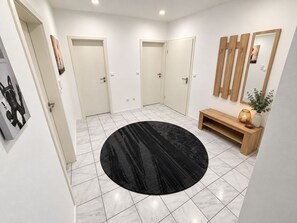 Ein Flur mit weißen Fliesen, einem schwarzen runden Teppich, einem Holzkonsole mit einer Vase und einer Lampe sowie einem Spiegel an der Wand.