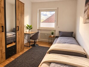 Ein Schlafzimmer mit einem Bett, einem Schreibtisch mit Stuhl, einem Fenster mit Blick auf ein Gebäude und einer Heizung.