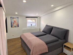 Een slaapkamer met een bed, een nachtkastje, een raam en een schilderij aan de muur.