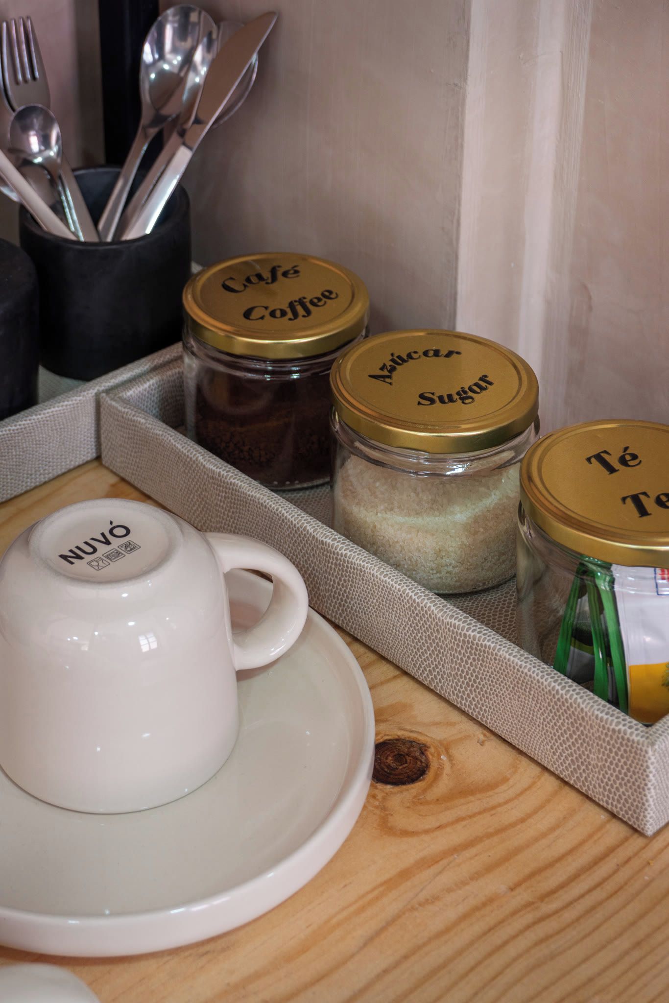 A kitchen counter with a white coffee cup, a tray with three glass jars labeled 'Cafe Coffee,' 'Azucar Sugar,' and 'Te,' and a black utensil holder.
