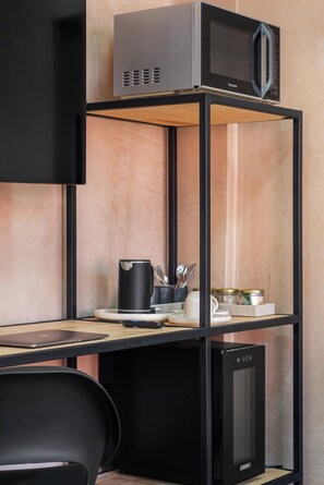 Una kitchenette con un microondas en un estante metálico negro, una cafetera, utensilios y una cocedora de arroz sobre una encimera de madera.