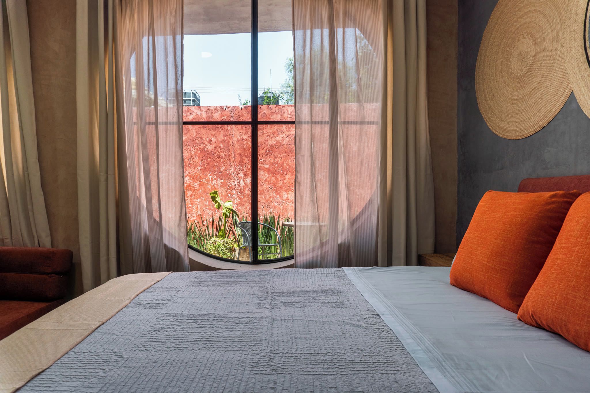 Une chambre avec un grand lit, une vue sur un mur rouge et sur la verdure à l’extérieur, et un objet rond en fibres tressées accroché au mur.