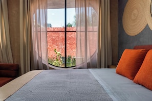 A bedroom with a large bed, a view of a red wall and greenery outside, and a round woven item on the wall.
