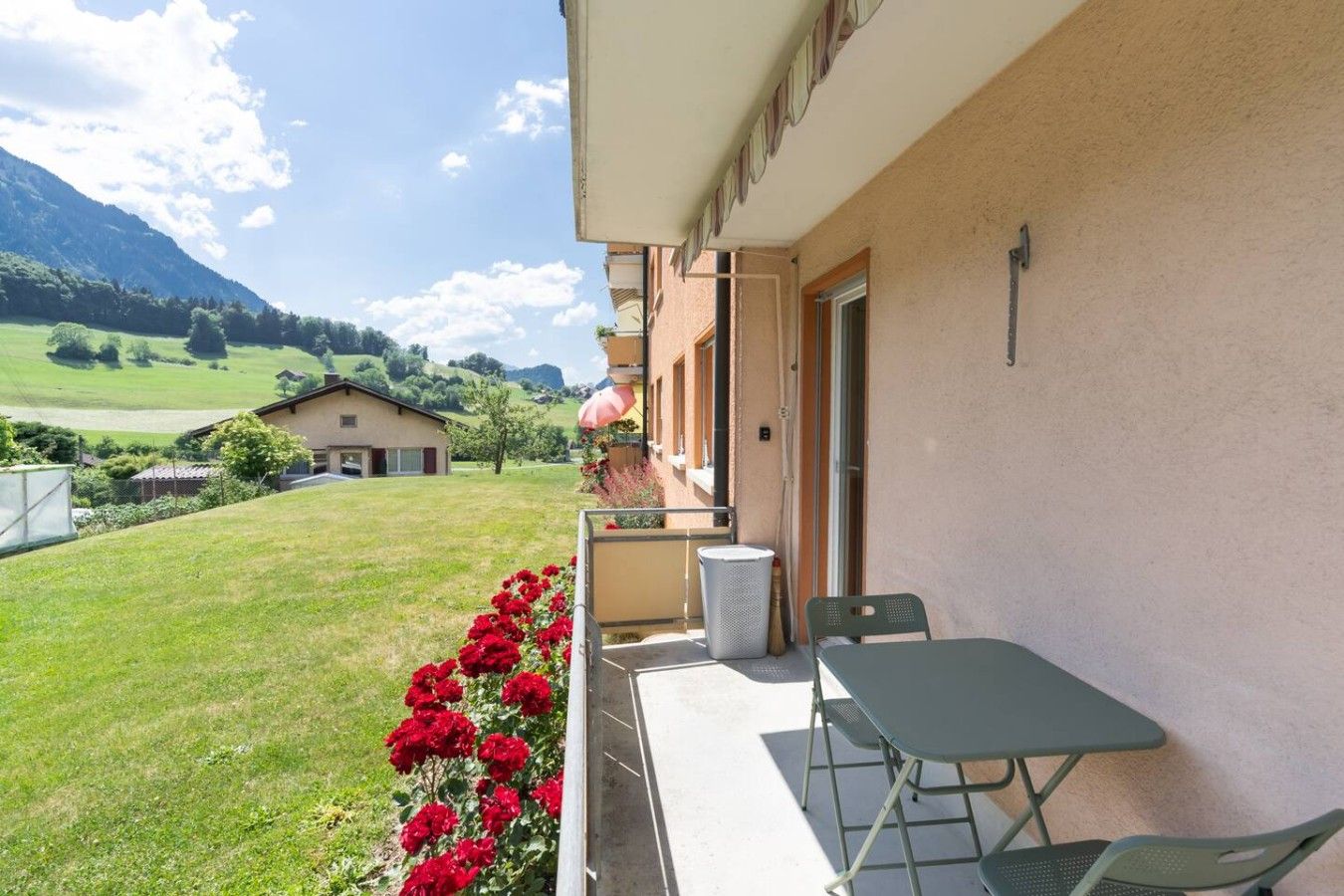 Ein Balkon mit Tisch und Stühlen, Blick auf eine grüne Landschaft und ein Gebäude mit hellbeiger Fassade.