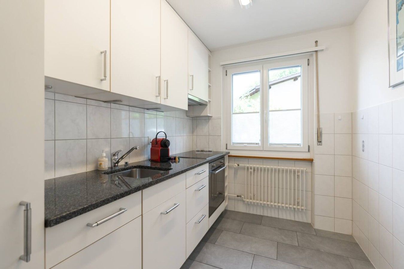 Eine Küche mit weißen Schränken, einer schwarzen Arbeitsplatte, einem roten Wasserkocher und einem Fenster mit Jalousien.