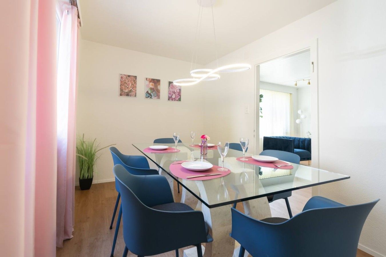 Een moderne eethoek met een glazen tafel, blauwe stoelen, roze placemats en een hanglamp.