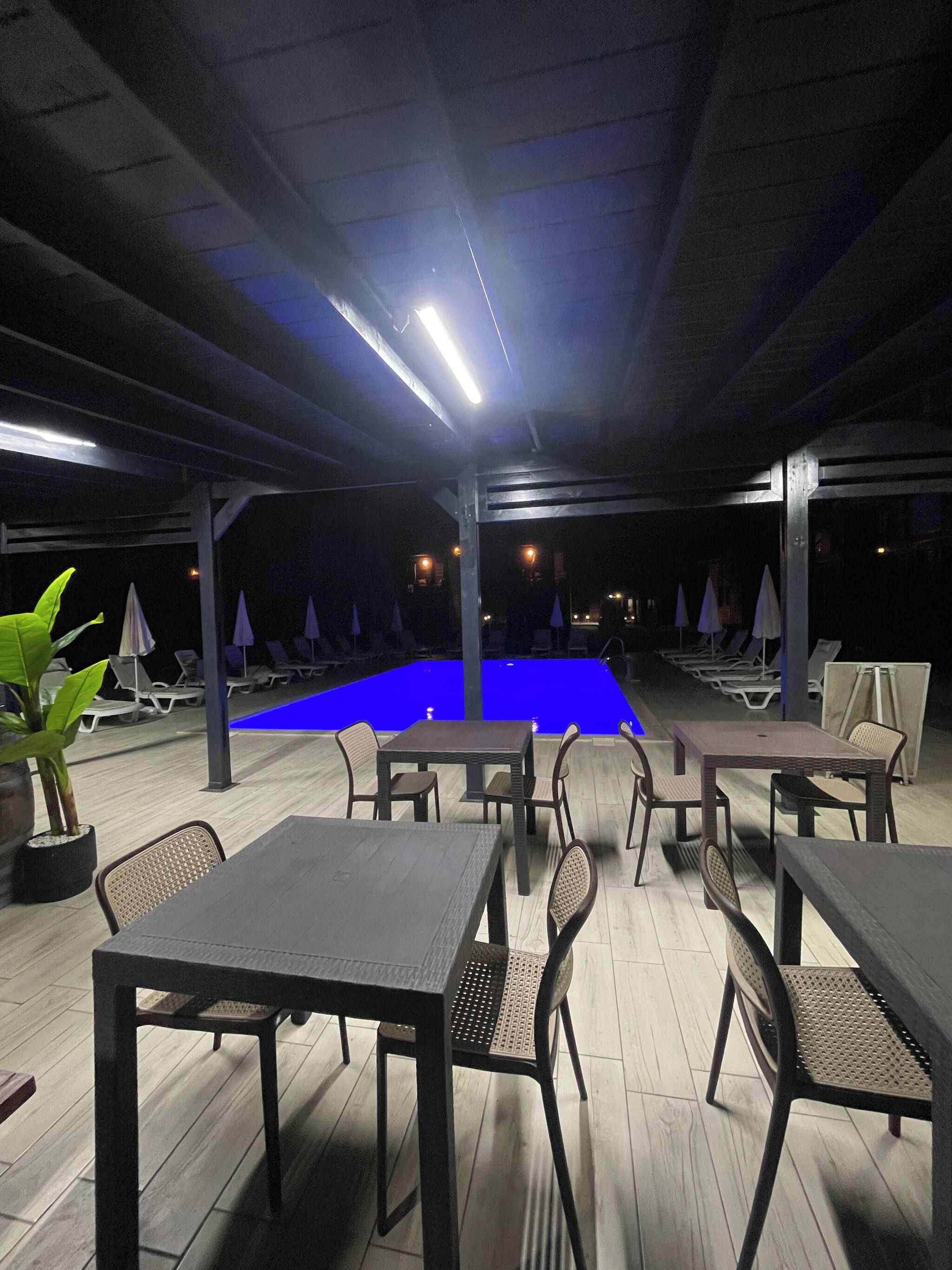 An outdoor pool area with lounge chairs and tables, surrounded by a covered patio with a wooden floor.