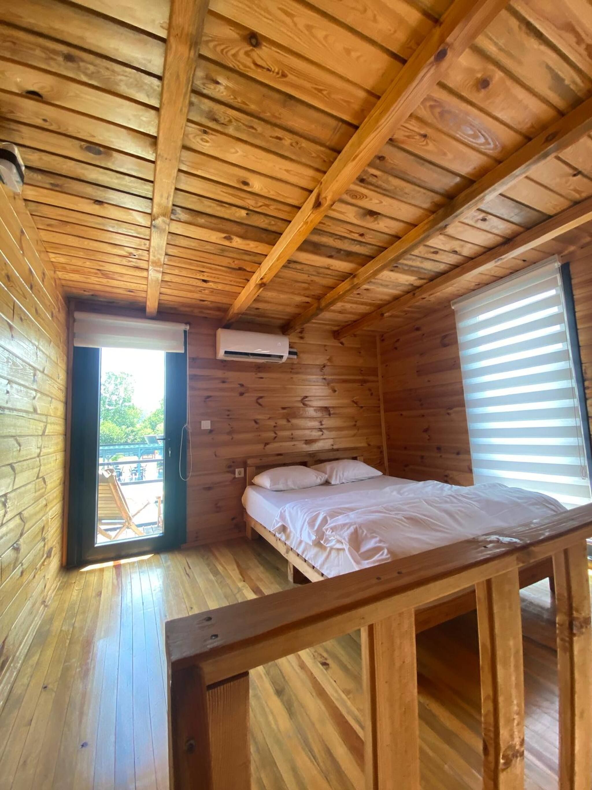 A wooden cabin room with a bed, an air conditioning unit, and a window with blinds.