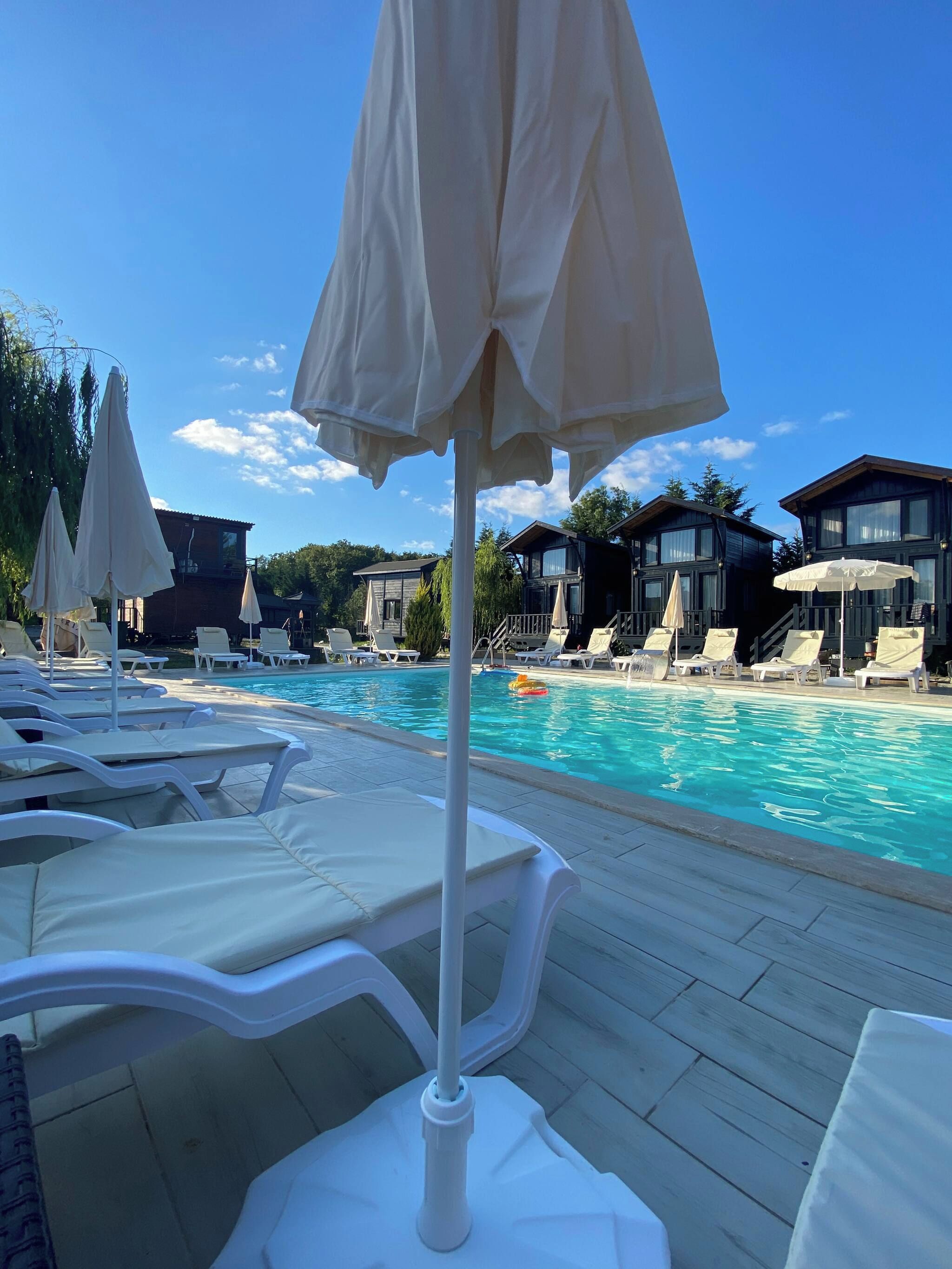 A poolside area with lounge chairs and umbrellas, a clear blue sky, and wooden buildings in the background.