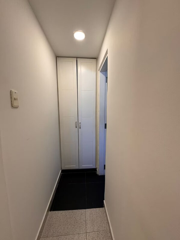 Un couloir étroit, avec un sol carrelé de carreaux noirs, des murs blancs et une porte blanche au fond.
