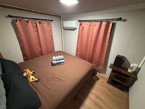 A bedroom with a bed, curtains, a small table with a speaker, and a wall-mounted air conditioner.