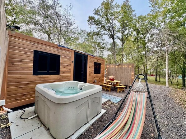 A wooden cabin with a hot tub, a hammock, and a picnic table in a wooded area.