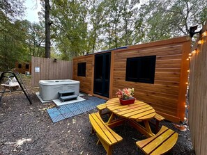A wooden cabin with a hot tub, picnic table, and a fenced area with string lights.