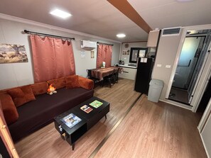 A living room with a sofa, a coffee table with magazines, a dining area with a table and chairs, and a kitchen area with a refrigerator and a sink.