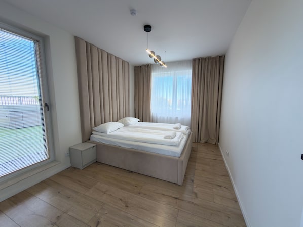 A modern bedroom with a large bed, a bedside table, a ceiling light fixture, and a view of the outside through a window with blinds.