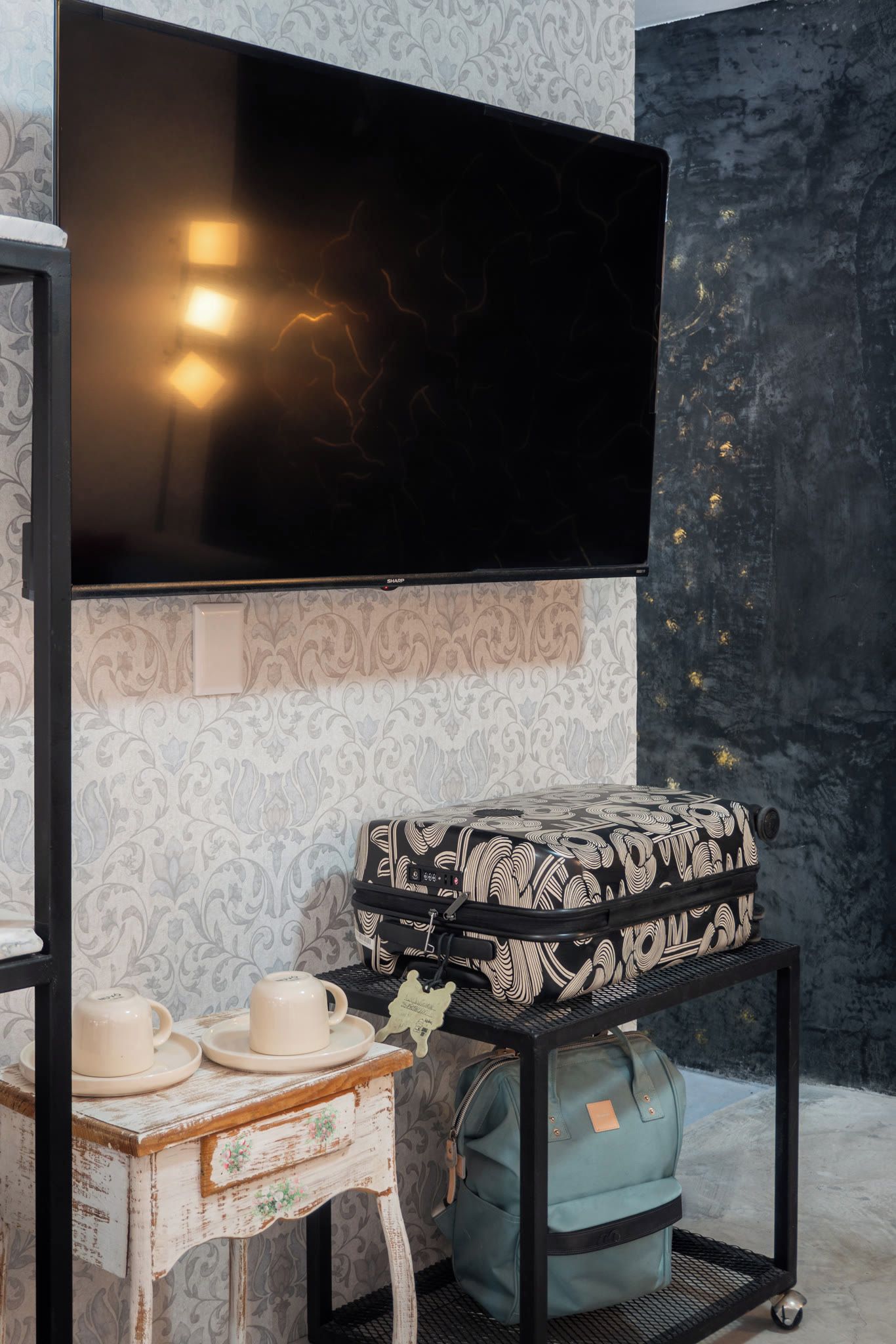 Una stanza con una TV a schermo piatto montata a parete, una valigia decorata su uno scaffale, un tavolino con tazza e piattino, e una borsa su un carrello.