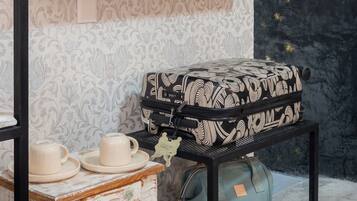 Una habitación con una televisión de pantalla plana montada en la pared, una maleta estampada en un estante, una mesita con una taza y un plato, y un bolso en un carrito.