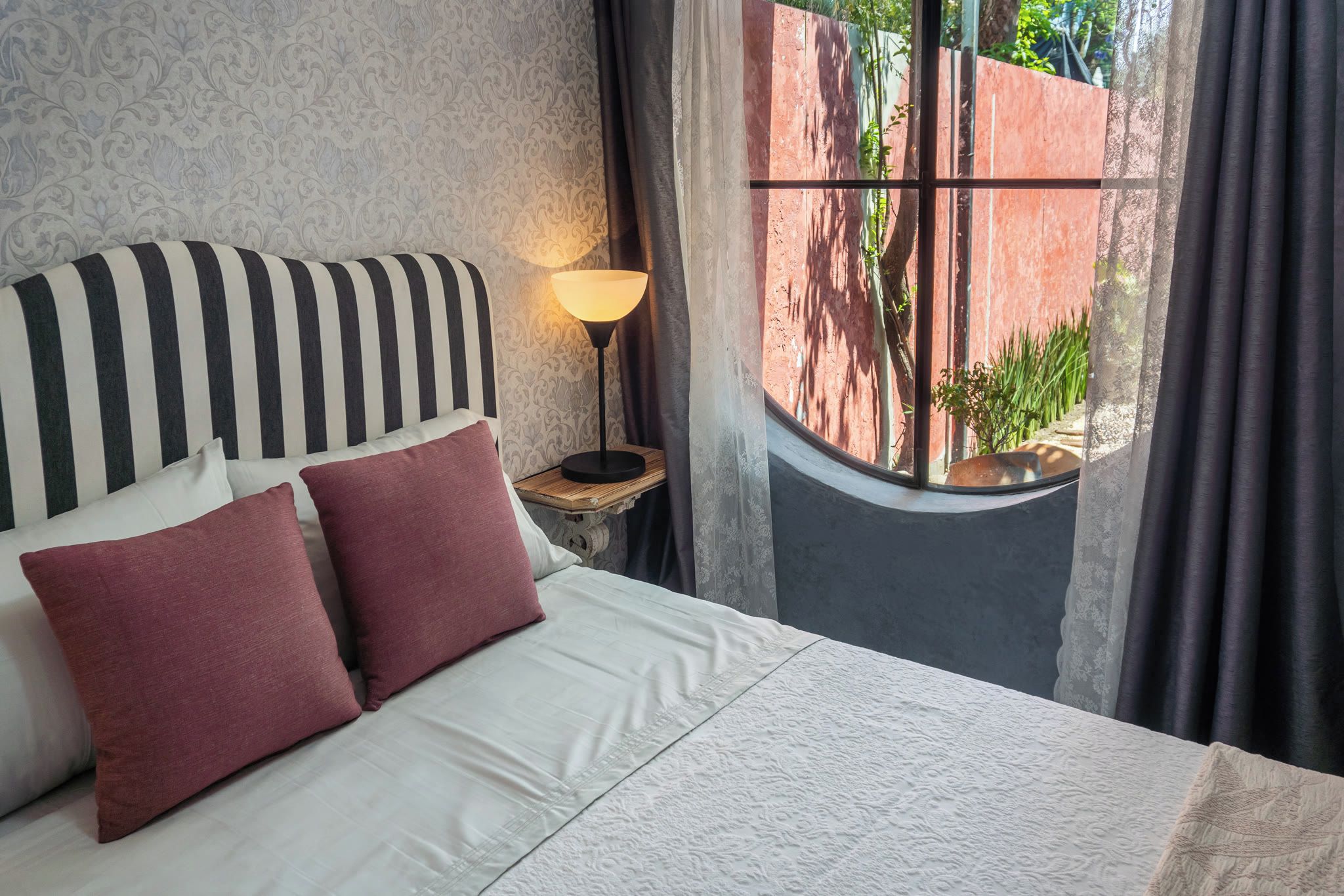 Ein Bett mit gestreifter Kopfteile, zwei Kissen, eine Nachttischlampe und ein Fenster mit Vorhängen.