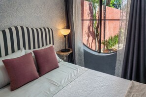 Una cama con cabecera a rayas, dos almohadas, una lámpara de noche y una ventana con cortinas.