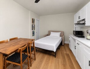 A compact room with a wooden dining table, a single bed, a kitchenette with a microwave, and a door with a small picture frame.