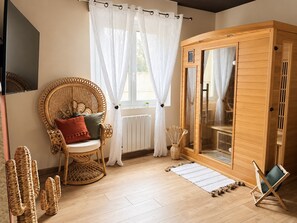 Ein Zimmer mit einem Korbsessel, einer Holzsauna und einem Fenster mit weißen Vorhängen.