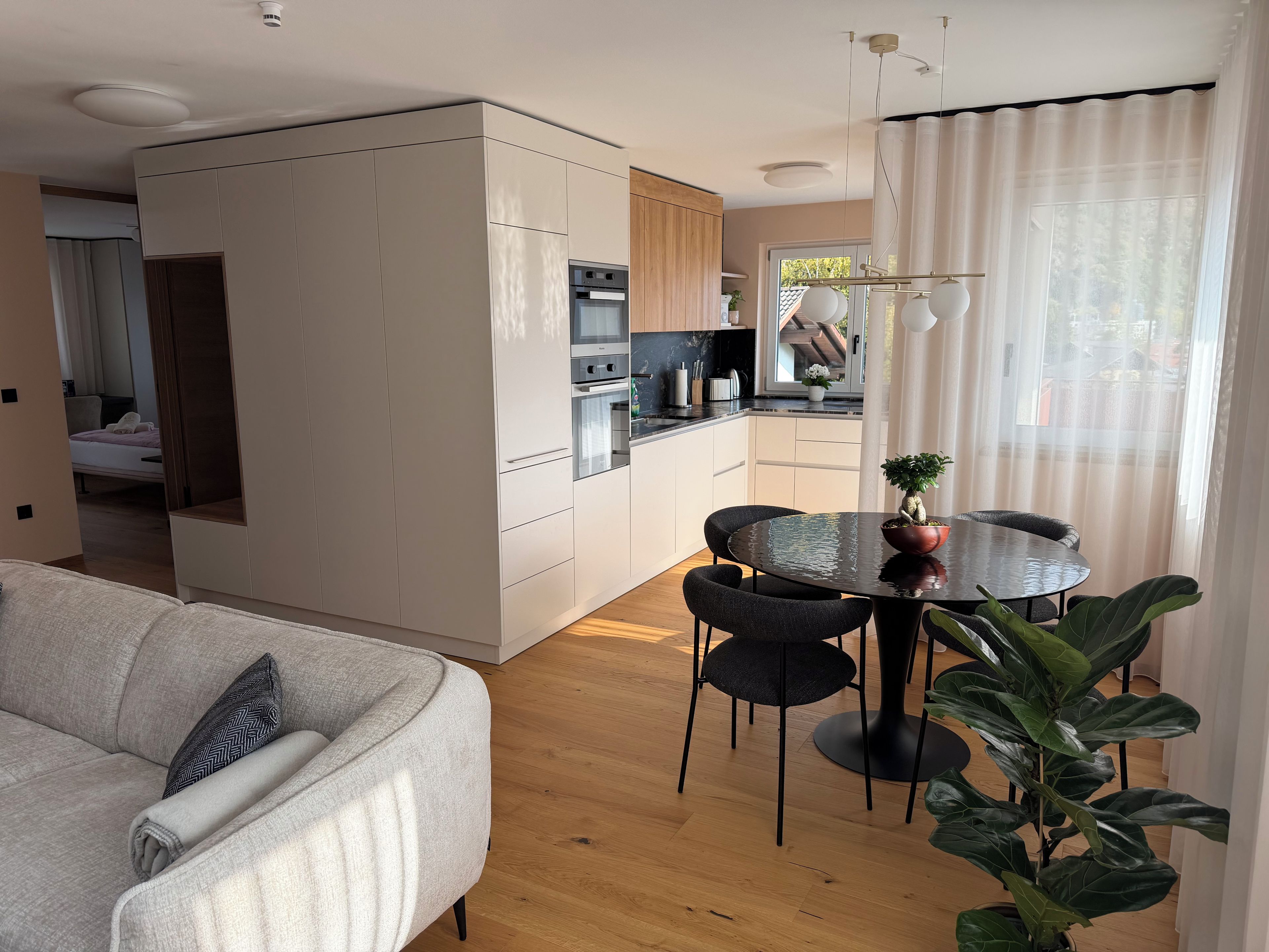 Una cocina moderna con electrodomésticos integrados, una zona de comedor con una mesa redonda y sillas, y una sala de estar con un sofá y una planta en maceta.