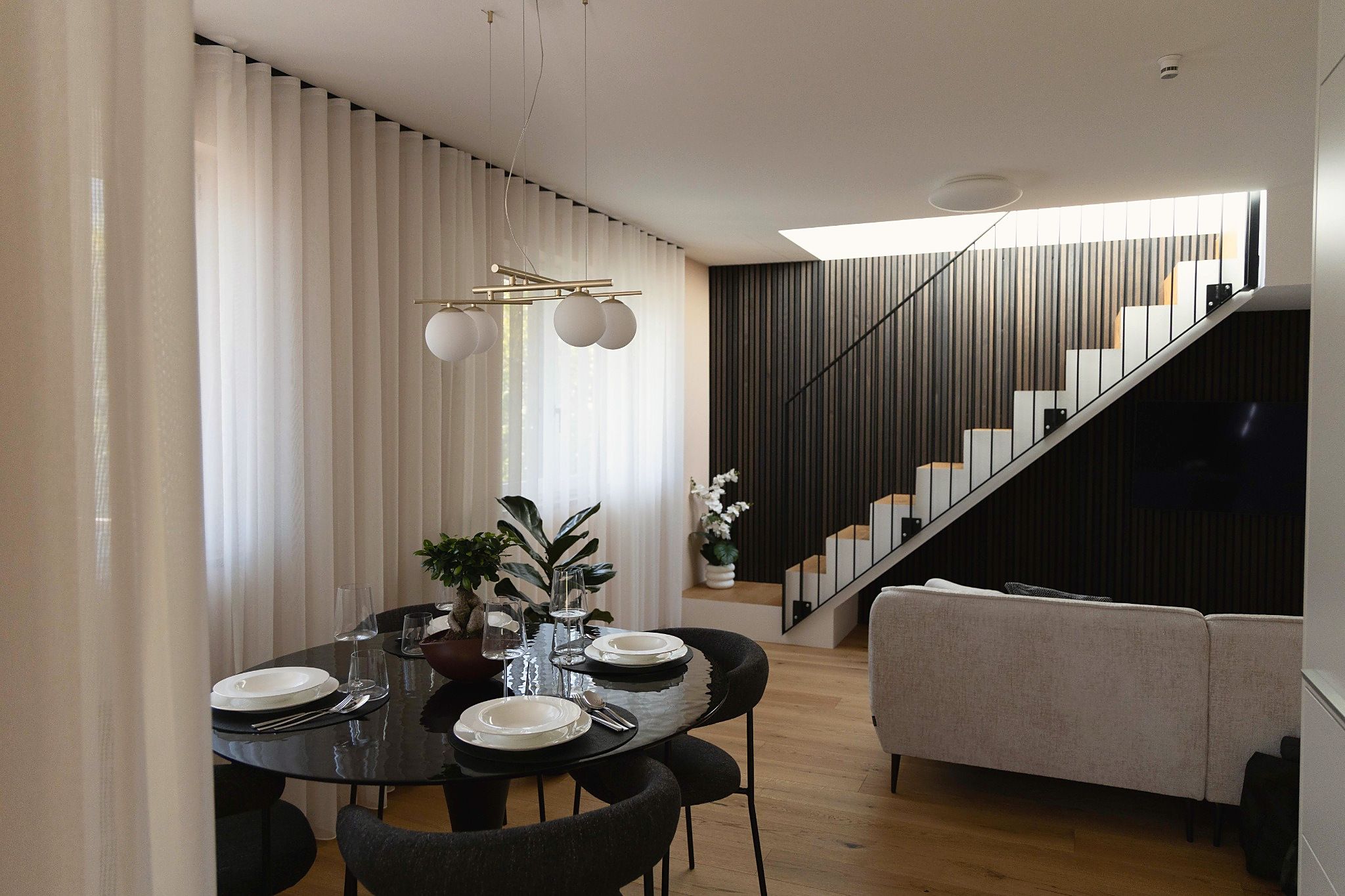 A modern dining area with a round table set for four, a staircase with a dark railing, and a light-colored sofa in the foreground.