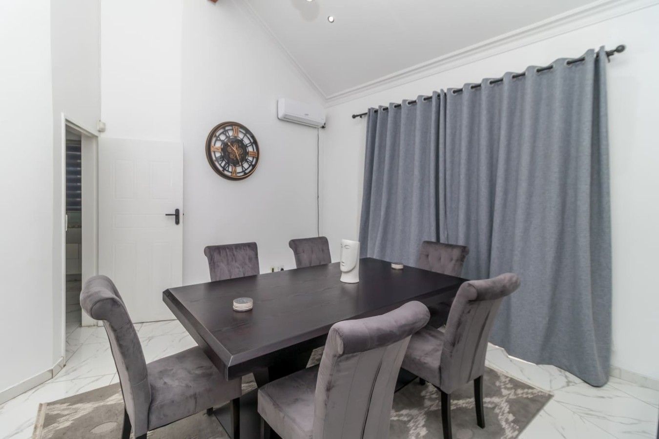Une salle de conférence avec une grande table en bois foncé, des chaises à revêtement gris, une horloge murale et des rideaux gris.