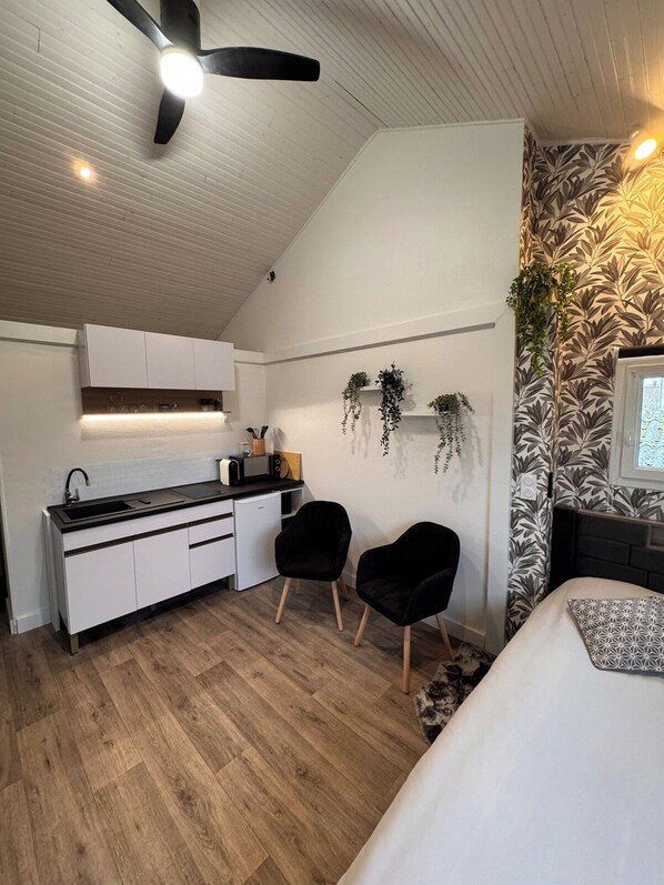 A compact kitchen with white cabinets, a black countertop, and a ceiling fan. There are two black chairs and a bed with a patterned pillow. - Brand-new design studio in Les Herbiers centre, covered parking (Les Herbiers)