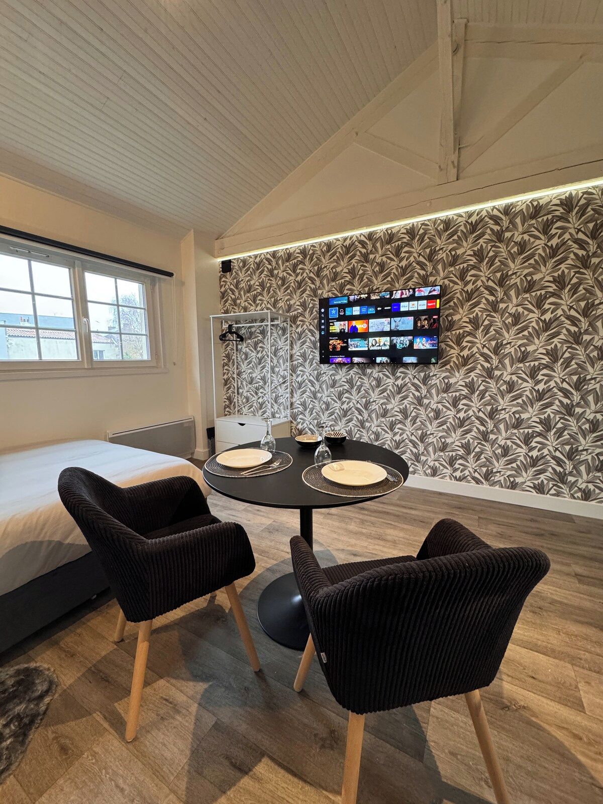 A modern room with a bed, a dining table with chairs, a flat-screen TV mounted on the wall, and a window with a view of buildings.