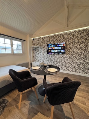 Una habitación moderna con cama, una mesa de comedor con sillas, un televisor de pantalla plana montado en la pared y un ventanal con vistas a edificios.