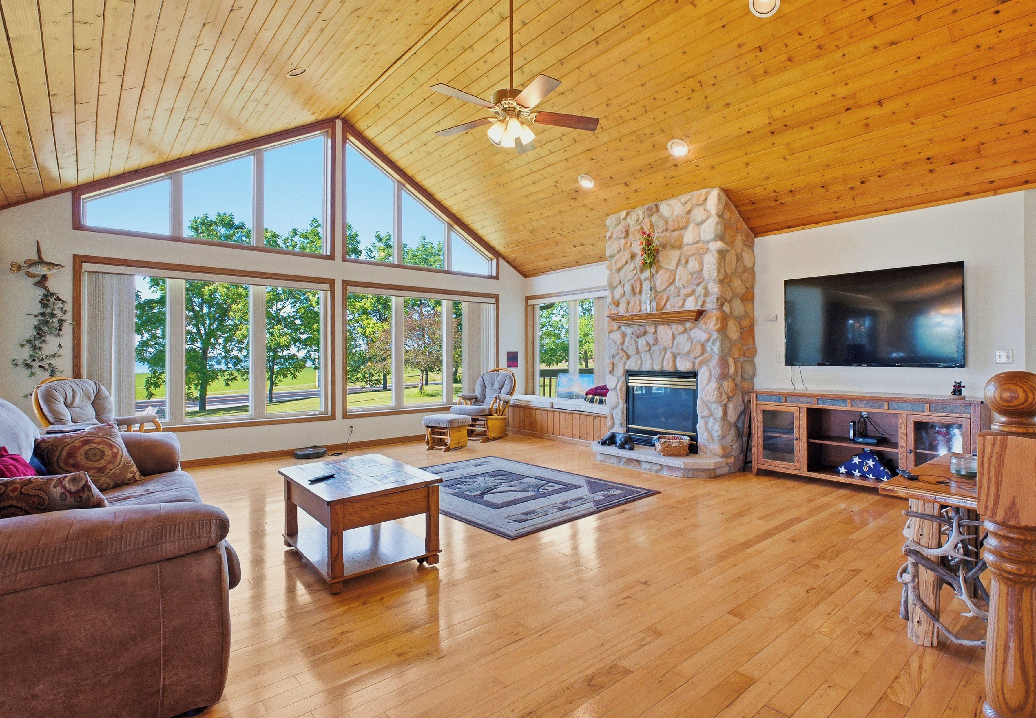 A spacious living room with a stone fireplace, large windows, and a ceiling fan.