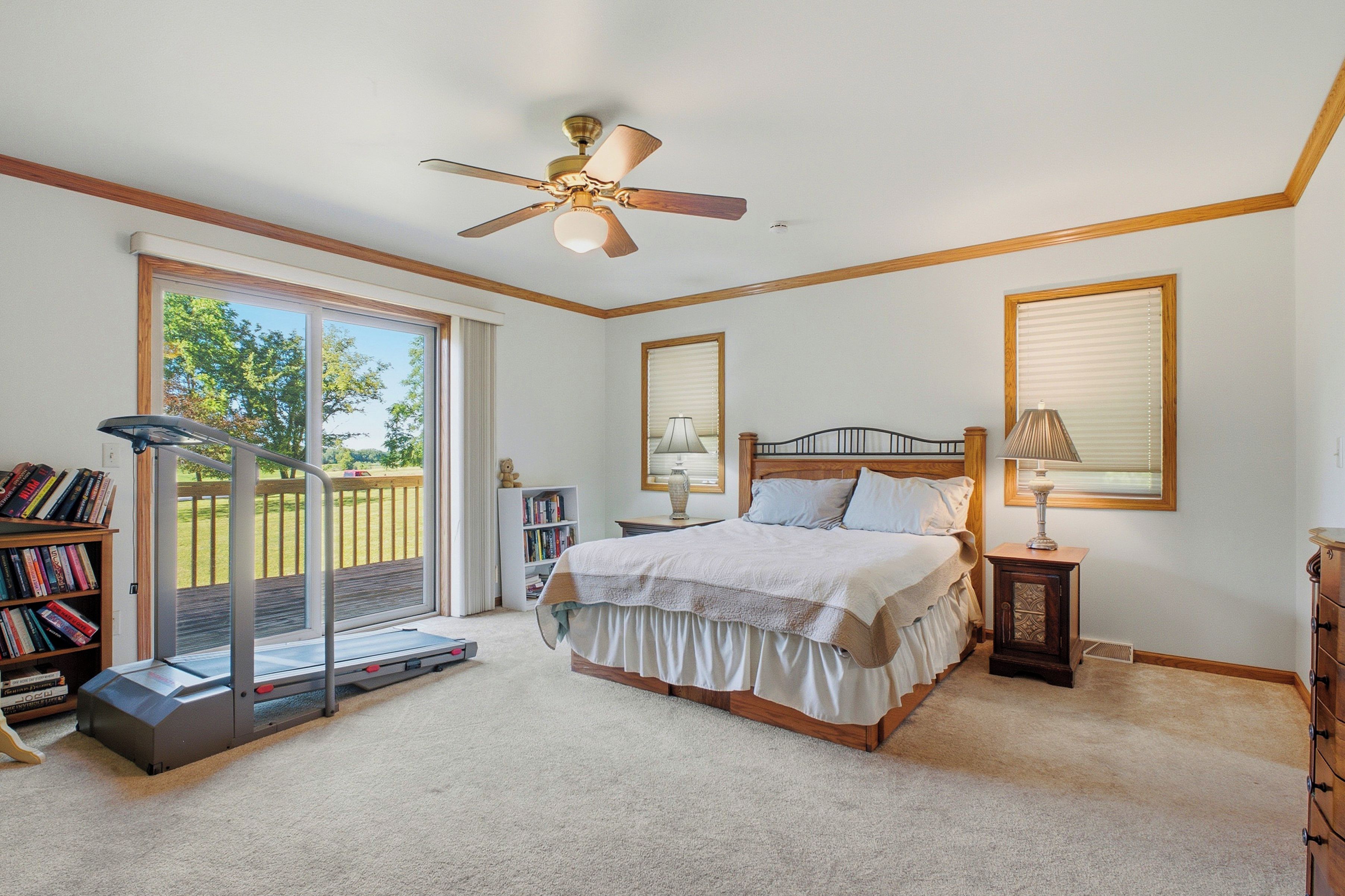 A bedroom with a bed, a treadmill, a bookshelf, and a window with a view of the outdoors.