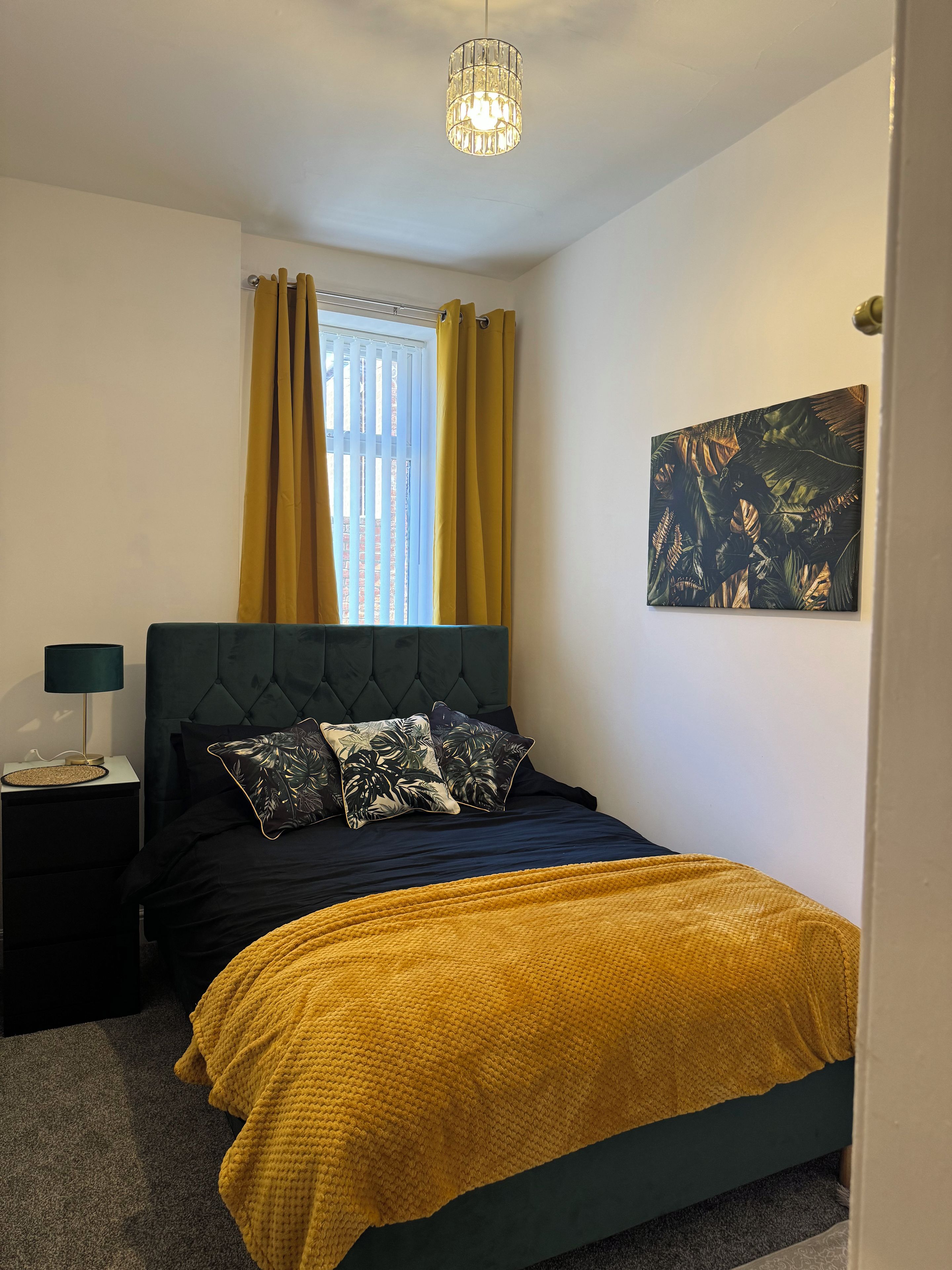 A bedroom with a bed, a nightstand, a lamp, a window with curtains, and a wall-mounted painting.