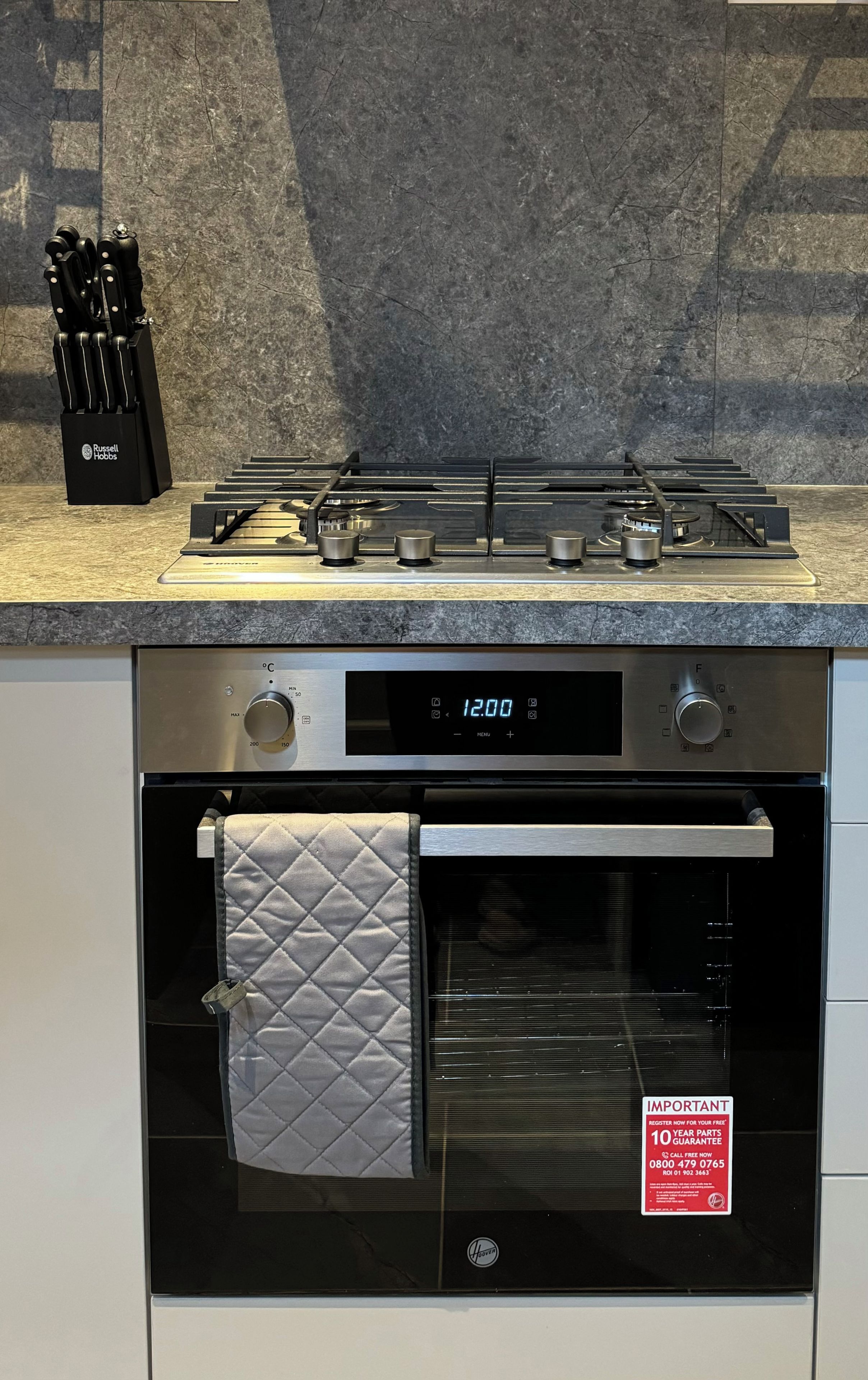 A kitchen with a digital clock displaying 12:00, a stove, oven, and a set of knives in a block.