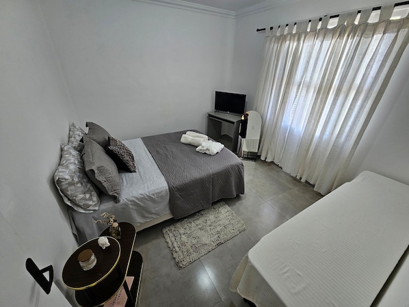 A bedroom with a bed, a desk, a TV, and a window with curtains.