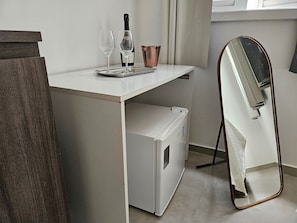 A compact room with a white desk, a mirror, a small refrigerator, and two wine glasses.