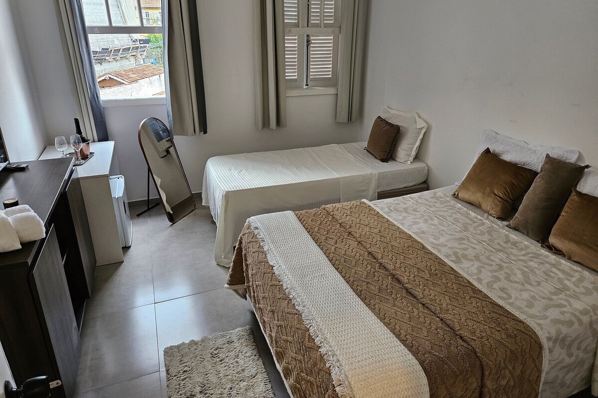 Una habitació d'hotel amb dos llits, un escriptori amb una cadira, una taula petita amb una ampolla i gots, i una finestra amb persianes.