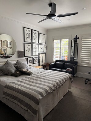 Ein Schlafzimmer mit einem Bett, einem Schreibtisch, einem Stuhl, einem Deckenventilator und einem Fenster mit Jalousien.