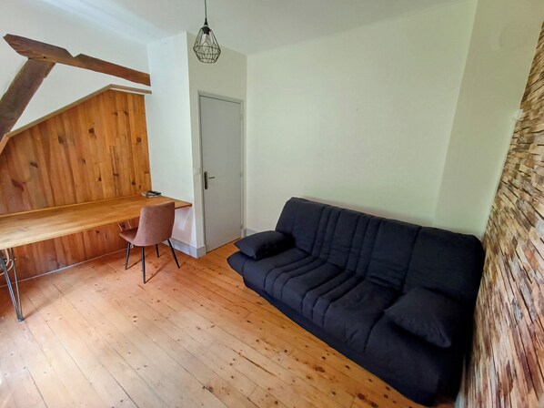 A room with a wooden floor, a black sofa, a wooden dining table with a chair, and a door. - New Listing (Charolles)