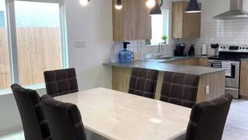 Una cocina moderna con una isla central, iluminación colgante y un área de comedor con una mesa de mármol y sillas tapizadas.