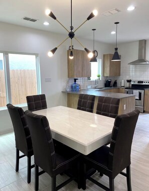 Una cocina moderna con una isla central, iluminación colgante y un área de comedor con una mesa de mármol y sillas tapizadas.
