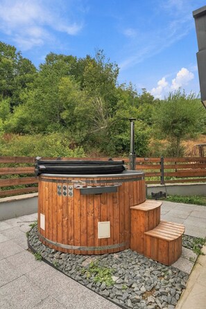 A wooden hot tub with a chimney, surrounded by a stone border and wooden steps, set against a backdrop of trees and a wooden fence. - Ren Villas Premium Mavrovo (Mavrovo)