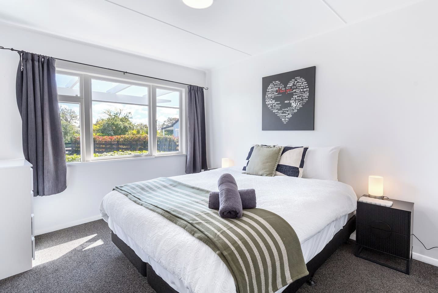 A hotel room with a large bed, a bedside table with a lamp, a window with curtains, and a heart-shaped artwork on the wall.