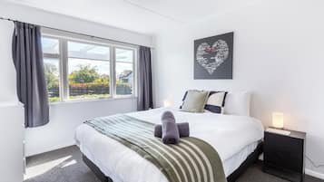 A hotel room with a large bed, a bedside table with a lamp, a window with curtains, and a heart-shaped artwork on the wall.